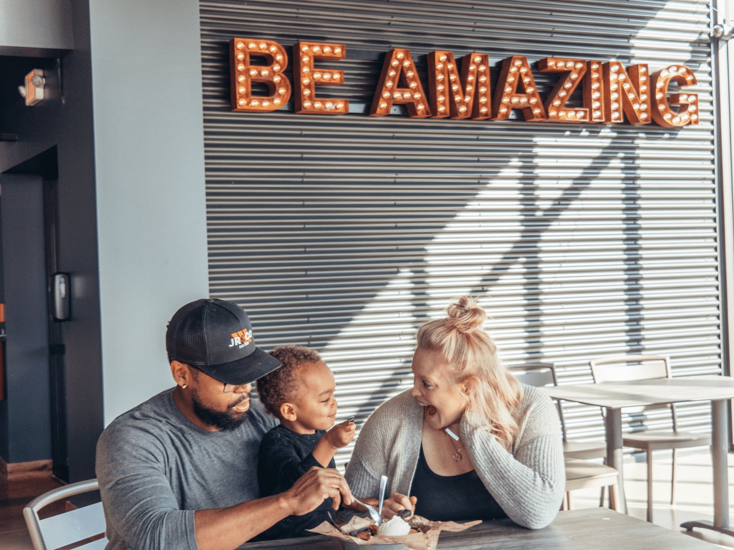 A man, child, and woman share dessert at a table under a lit sign that reads BE AMAZING in a modern cafe.