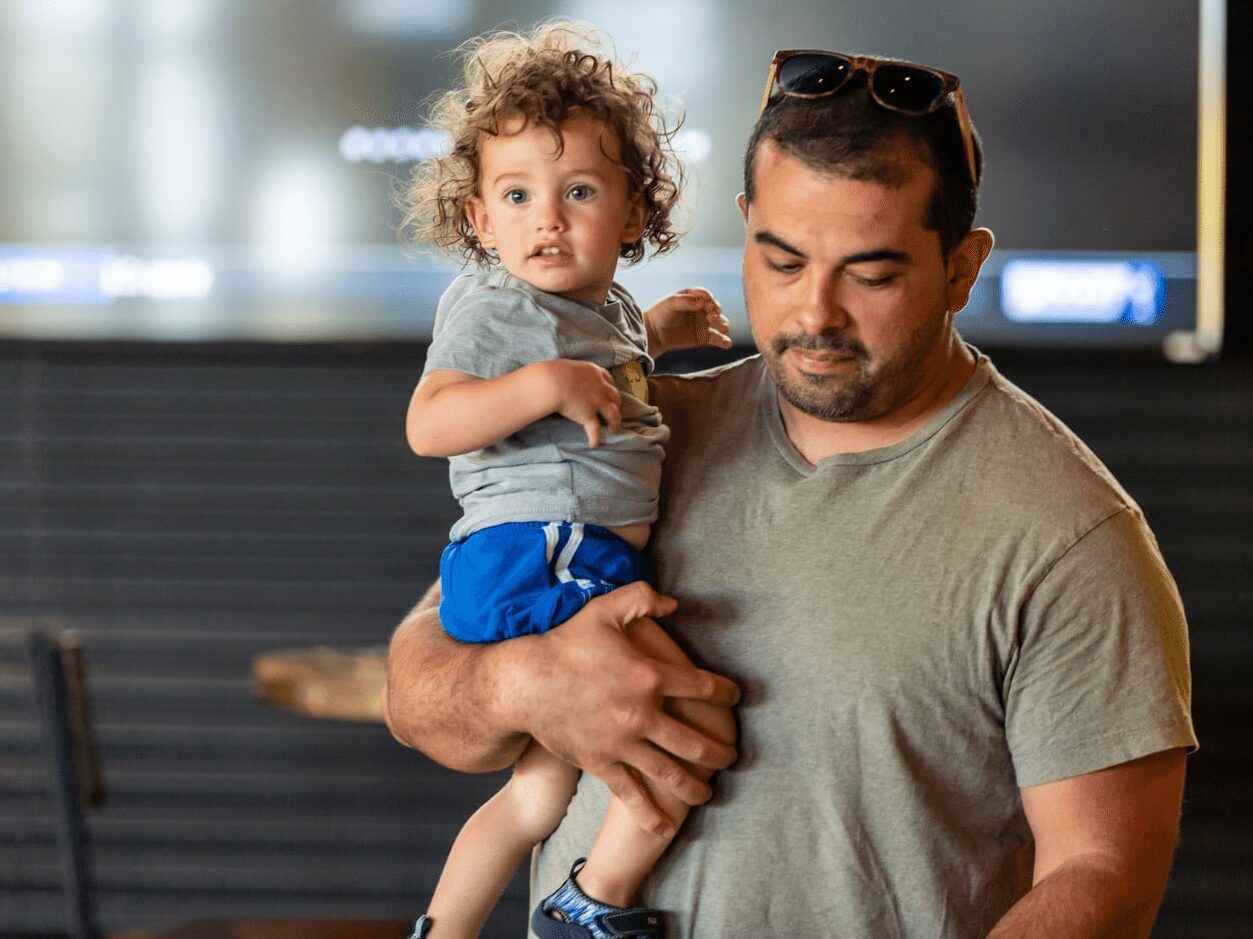 A man wearing sunglasses on his head holds a young child with curly hair, both looking in different directions, indoors with a blurred background.