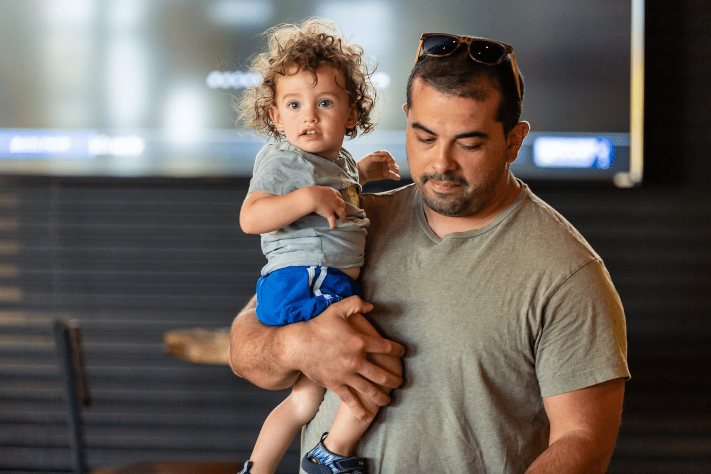 A man wearing sunglasses on his head holds a young child with curly hair, both looking in different directions, indoors with a blurred background.