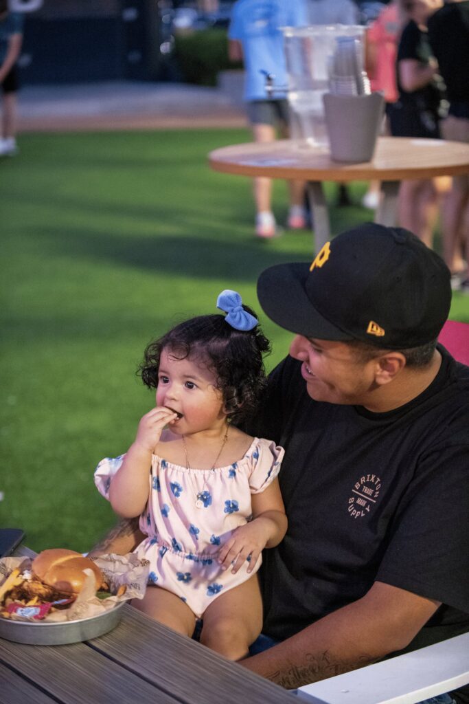 A man smiles at a young girl sitting on his lap as she eats food. They are outdoors at a table with burgers and fries, with people and green grass in the background.