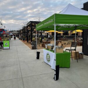 A green and white vendor tent with a table displaying products is set up on a wide outdoor sidewalk near picnic tables, string lights, and empty seating under a cloudy sky.