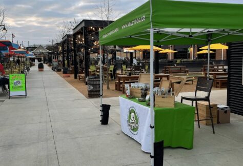 A green and white vendor tent with a table displaying products is set up on a wide outdoor sidewalk near picnic tables, string lights, and empty seating under a cloudy sky.