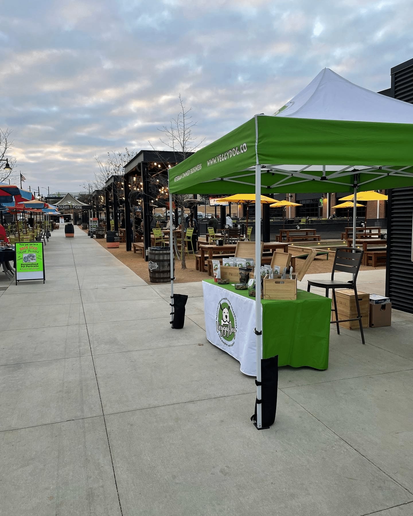 A green and white vendor tent with a table displaying products is set up on a wide outdoor sidewalk near picnic tables, string lights, and empty seating under a cloudy sky.
