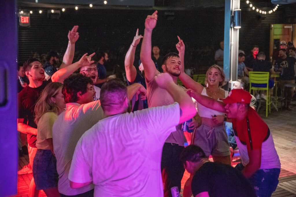 A group of people enthusiastically singing and dancing together under string lights at an indoor event, illuminated by colorful lighting, with others seated in the background.