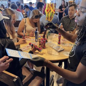 People sit and stand around a table playing bingo, holding cards and colorful daubers. The setting is casual and lively, with drinks and snacks on the table. Balloons and decorations are visible in the background.