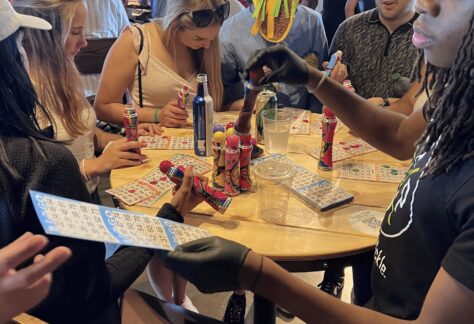 People sit and stand around a table playing bingo, holding cards and colorful daubers. The setting is casual and lively, with drinks and snacks on the table. Balloons and decorations are visible in the background.