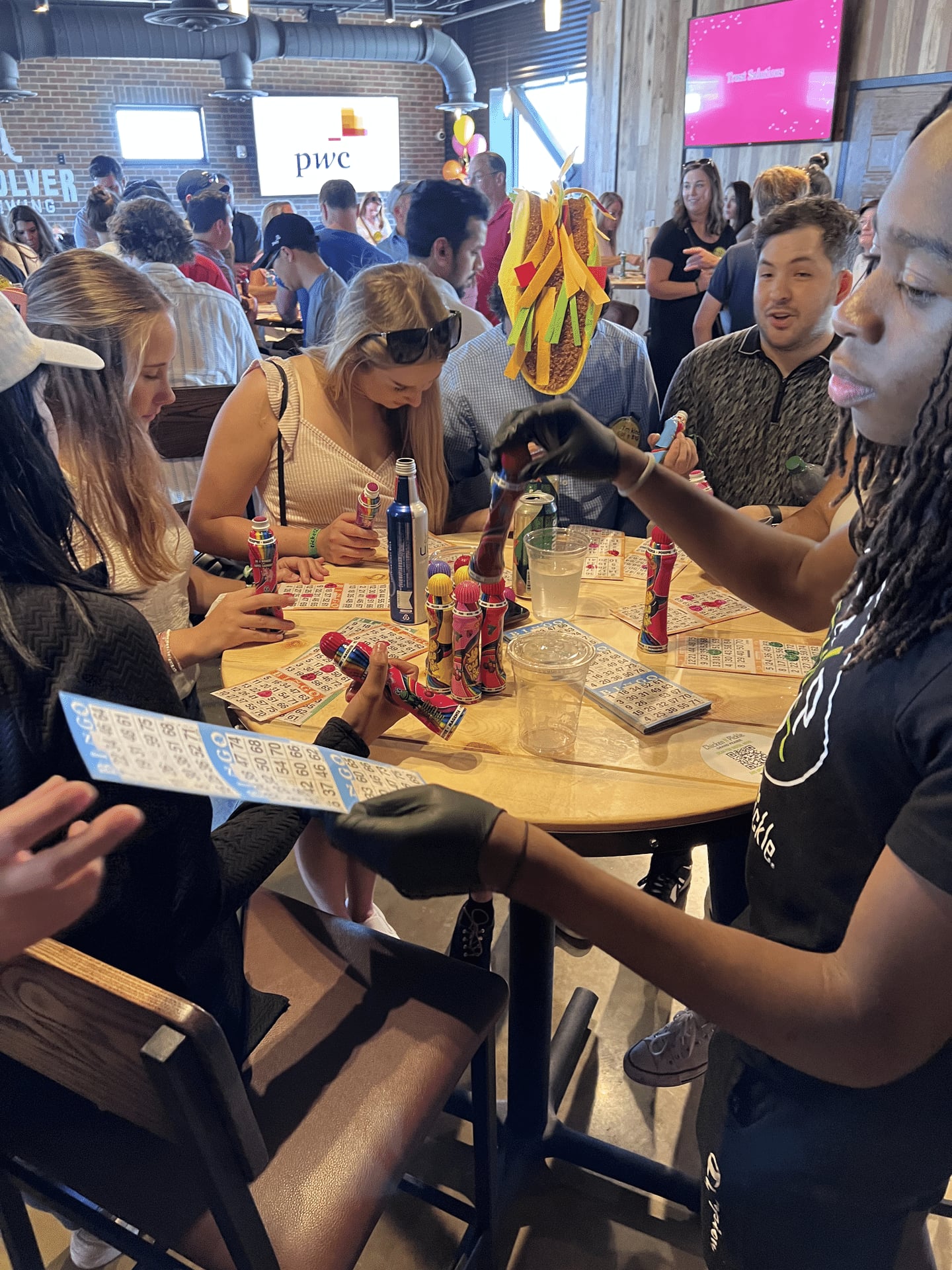 People sit and stand around a table playing bingo, holding cards and colorful daubers. The setting is casual and lively, with drinks and snacks on the table. Balloons and decorations are visible in the background.