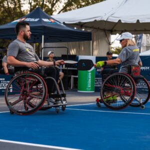 Two athletes in wheelchairs play pickleball on an outdoor court. Both wear gray shirts and hold paddles, with a net between them. Spectators and tents are visible in the background.