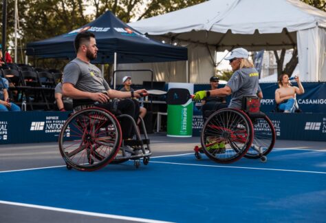 Two athletes in wheelchairs play pickleball on an outdoor court. Both wear gray shirts and hold paddles, with a net between them. Spectators and tents are visible in the background.