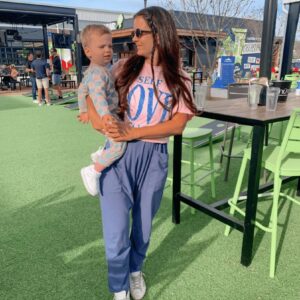 A woman wearing sunglasses holds a young child outdoors on artificial grass near green bar stools and tables during the Tomato Dinner at Chicken N Pickle, with other people and buildings in the background on a sunny day.