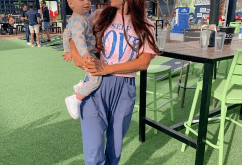 A woman wearing sunglasses holds a young child outdoors on artificial grass near green bar stools and tables during the Tomato Dinner at Chicken N Pickle, with other people and buildings in the background on a sunny day.