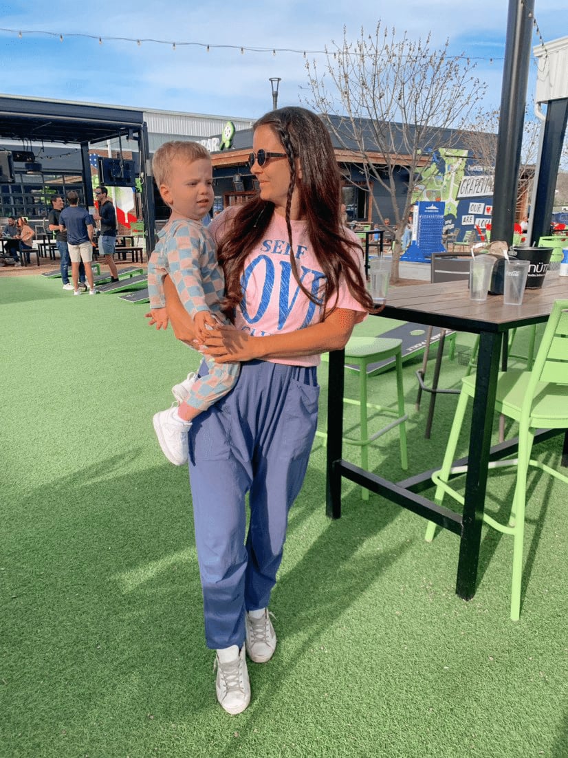 A woman wearing sunglasses holds a young child outdoors on artificial grass near green bar stools and tables during the Tomato Dinner at Chicken N Pickle, with other people and buildings in the background on a sunny day.