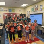 A classroom of young children stand smiling, each holding up a red book. Several adults stand behind them. The classroom is colorful with posters, desks, and an interactive screen.