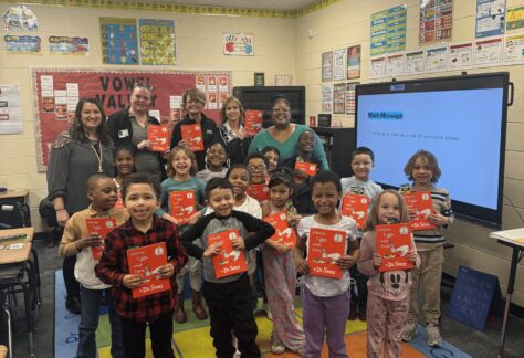 A classroom of young children stand smiling, each holding up a red book. Several adults stand behind them. The classroom is colorful with posters, desks, and an interactive screen.
