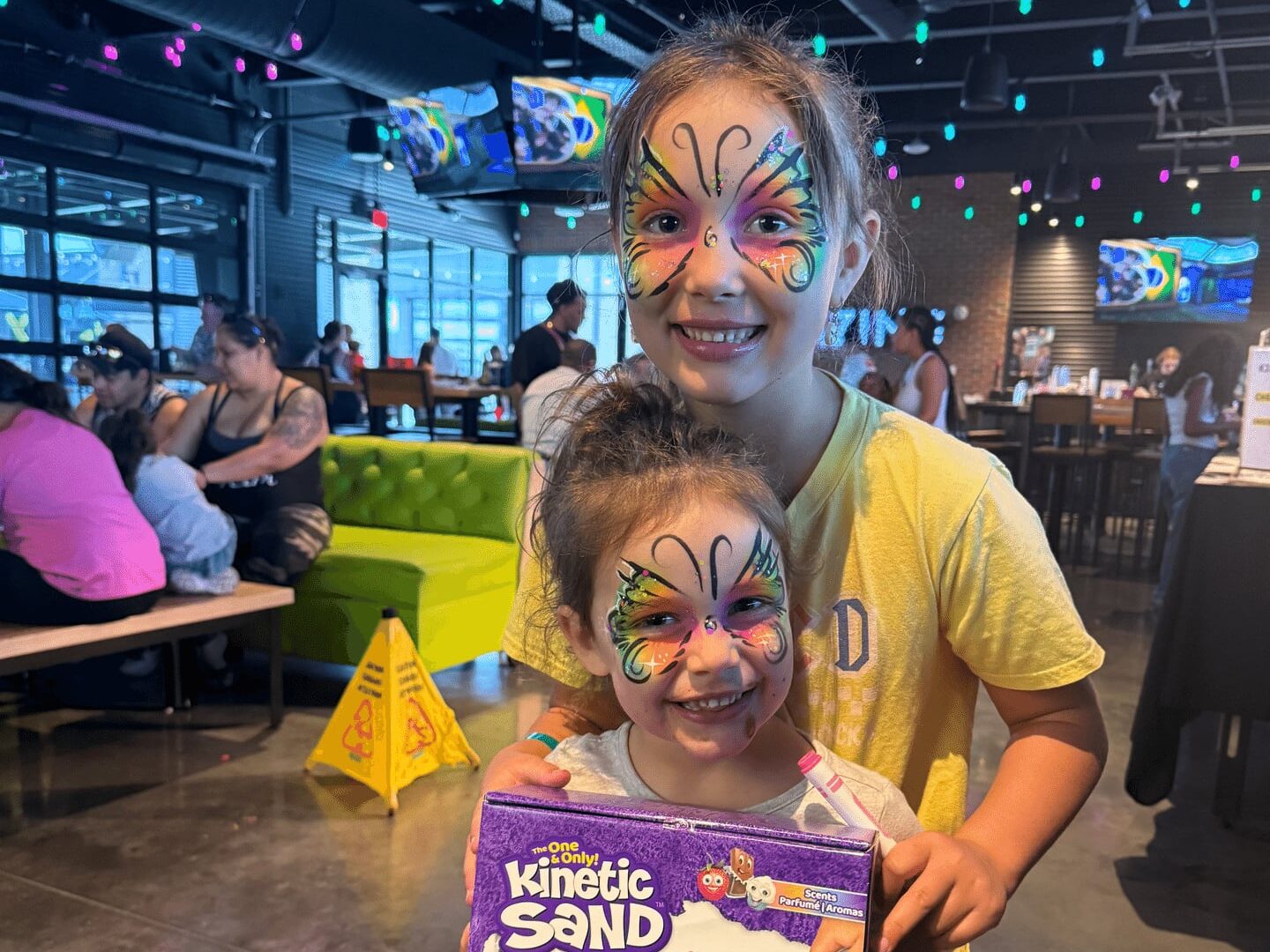 Two smiling children with butterfly face paint stand inside a lively, colorful restaurant. The younger child holds a Kinetic Sand Ice Cream Treats box. People sit at tables in the background under string lights.