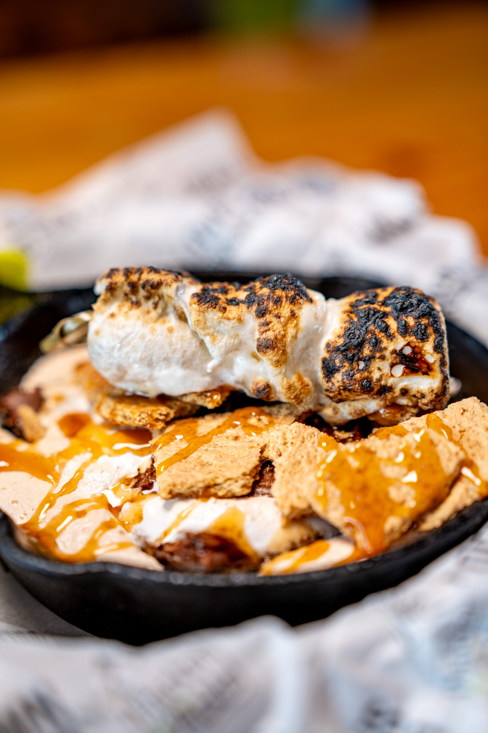 A close-up of a skillet dessert with toasted marshmallows, graham cracker pieces, melted chocolate, and caramel drizzle, resembling a smore.