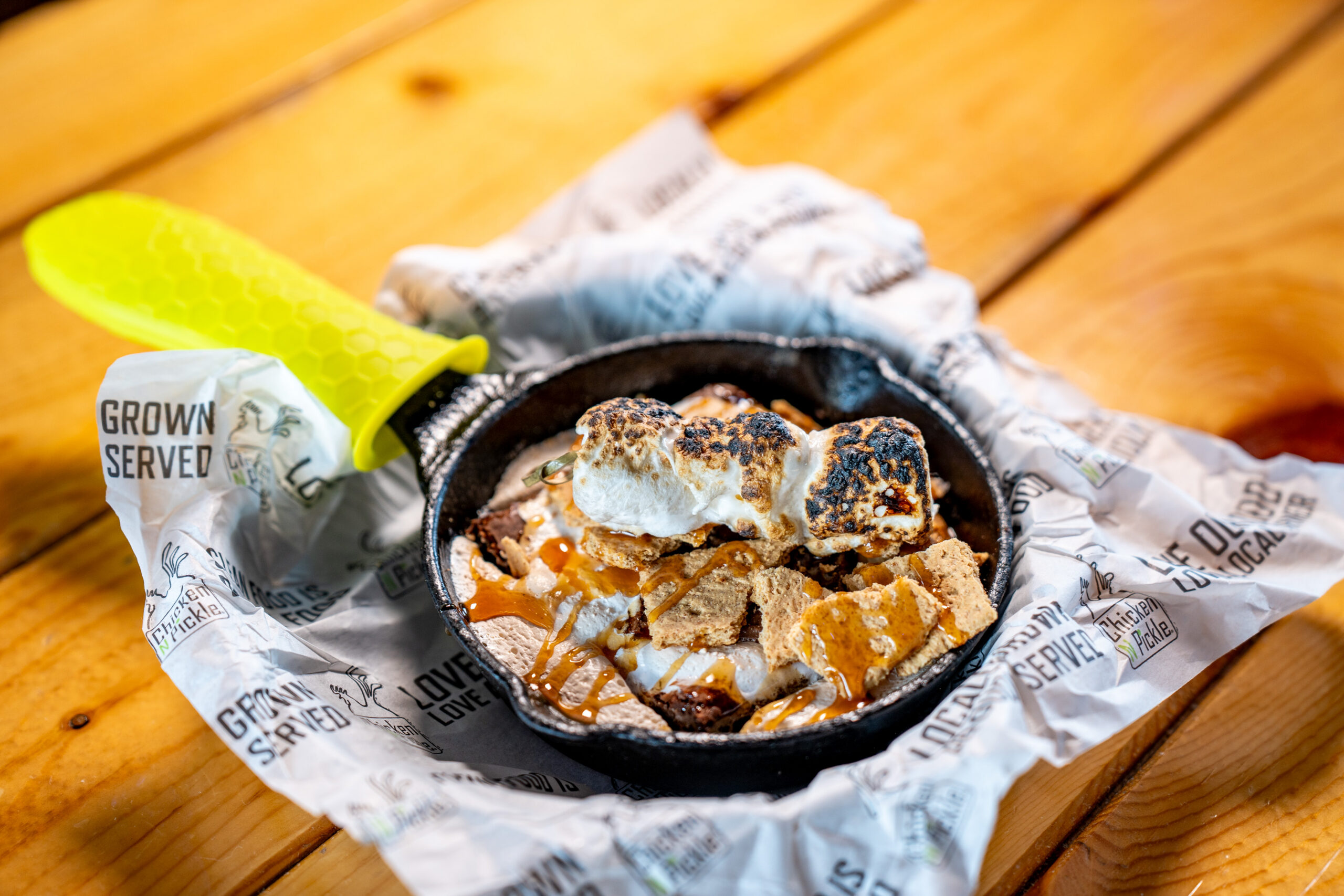 A small cast iron skillet with toasted marshmallows, graham crackers, and chocolate, served on branded paper atop a wooden table. A bright green handle cover is on the skillet.