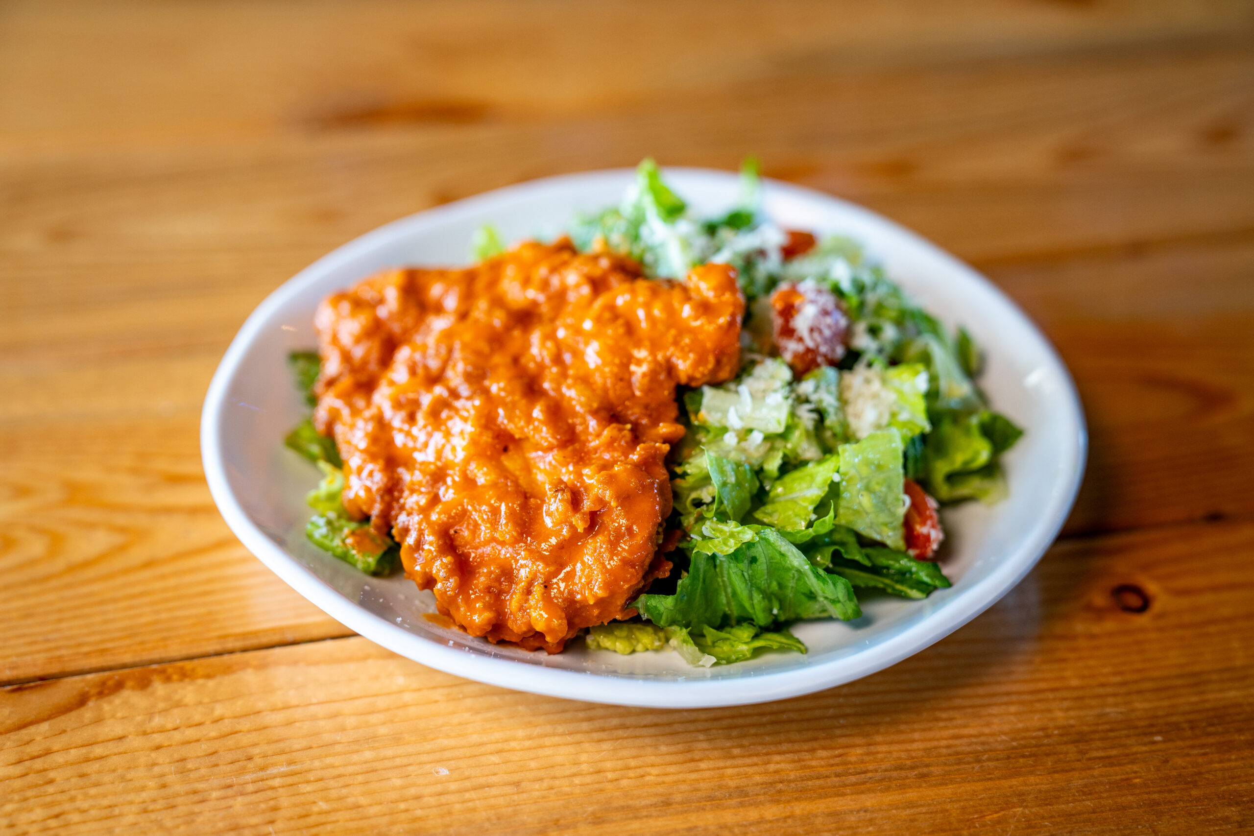 A white plate with buffalo chicken in orange sauce next to a green salad with lettuce, tomatoes, and a creamy dressing, on a wooden table.
