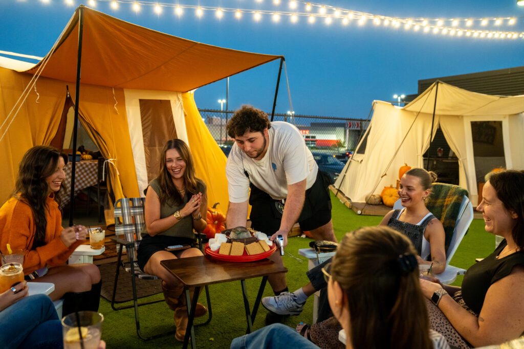 A group of people laugh and enjoy snacks together at a cozy outdoor campsite at dusk, sitting around a table with tents and string lights in the background.
