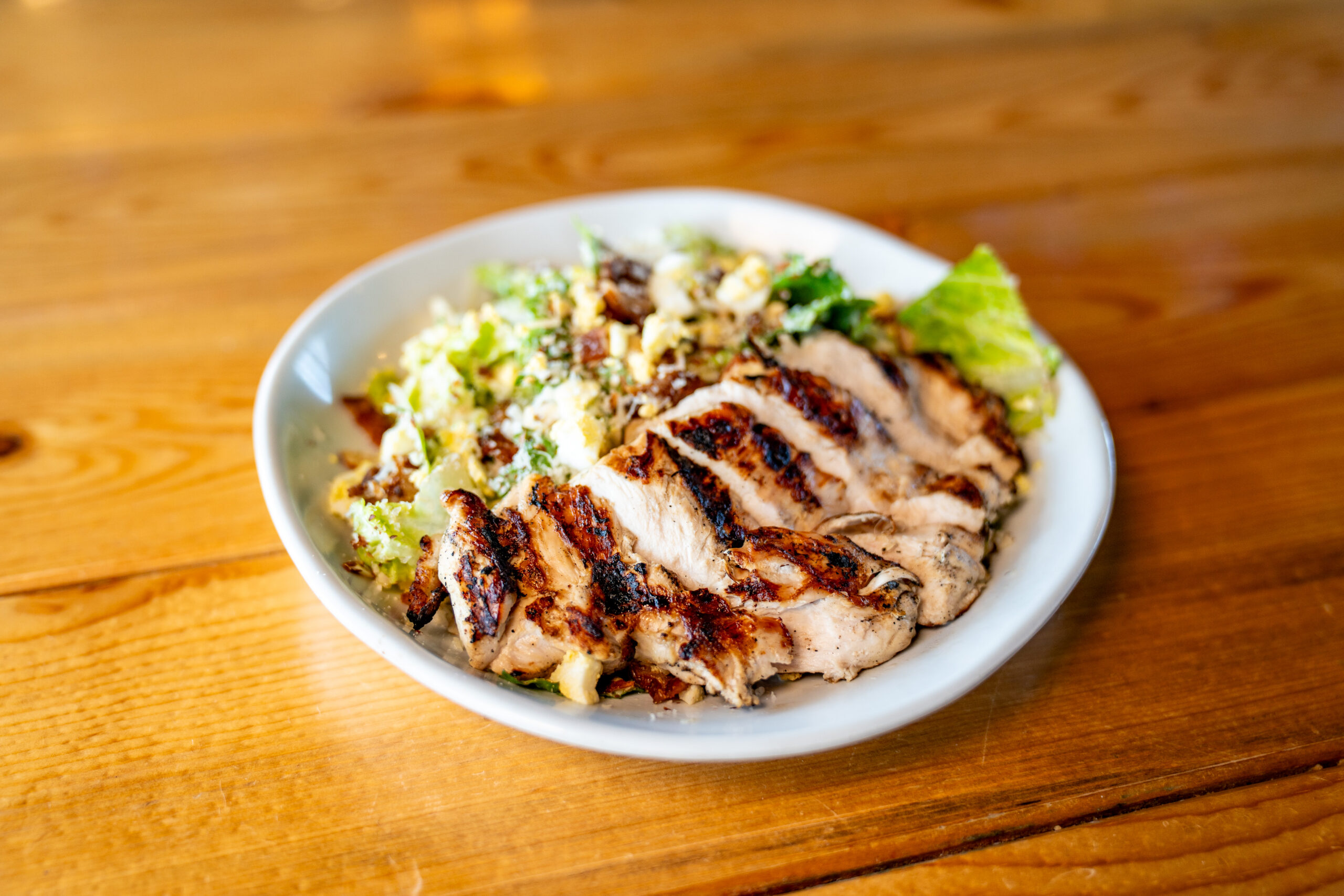 A white bowl filled with grilled, sliced chicken breast on top of a green salad with chopped nuts and cheese, set on a wooden table.