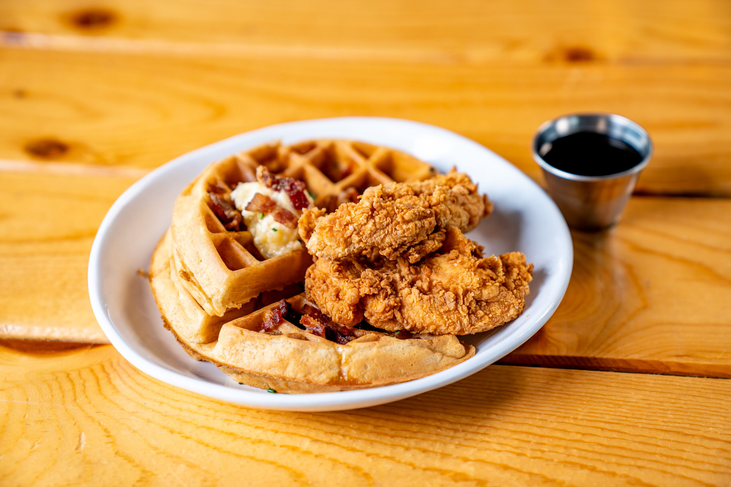 A white plate with two pieces of fried chicken on top of two waffle quarters, served with a small metal cup of syrup, on a wooden table.