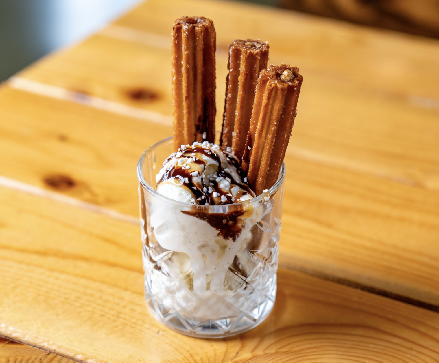 A glass filled with vanilla ice cream, topped with chocolate sauce, sprinkles, and four churros, sitting on a wooden table.
