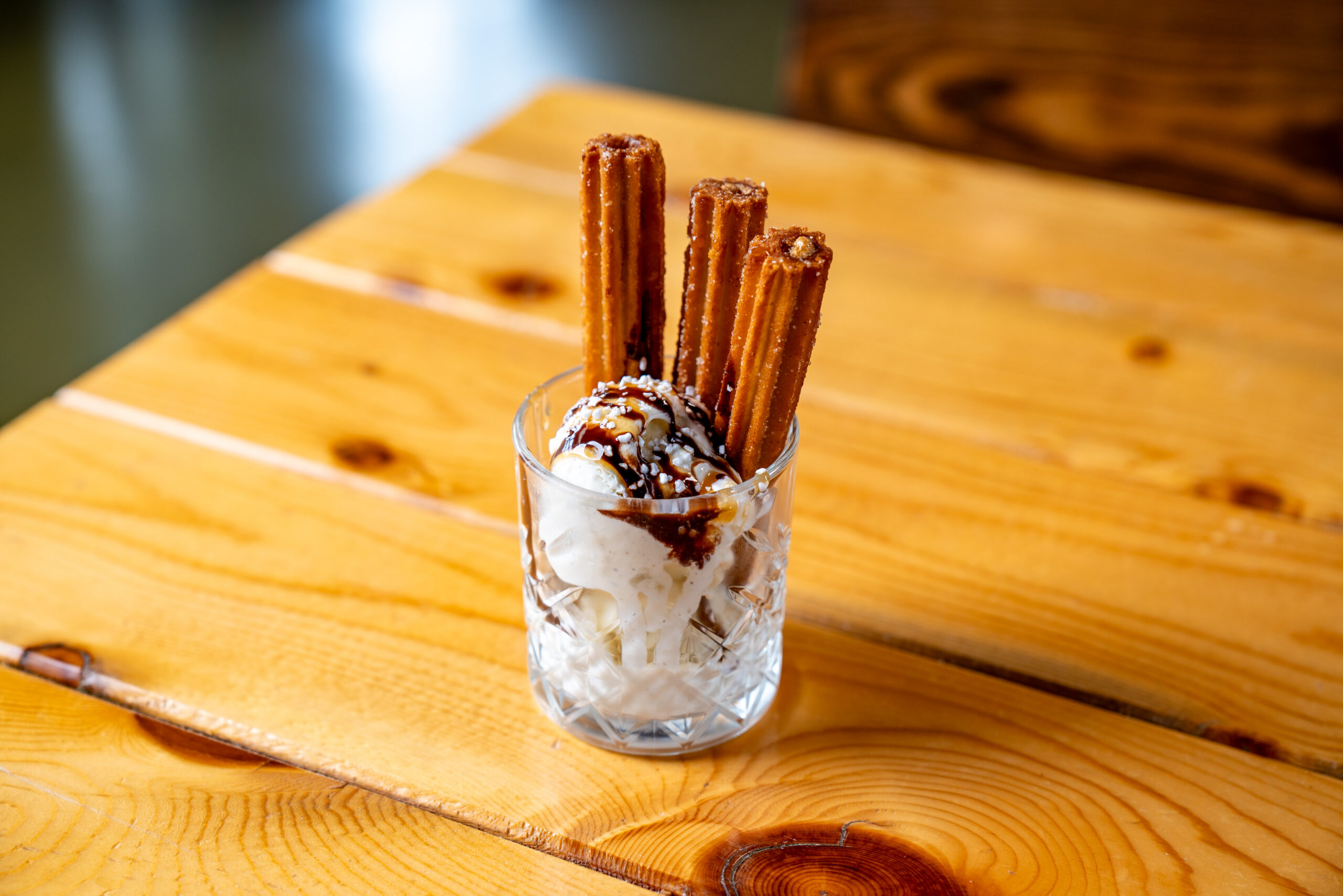 A glass cup filled with vanilla ice cream, topped with chocolate sauce, sprinkles, and three churros, sits on a wooden table.