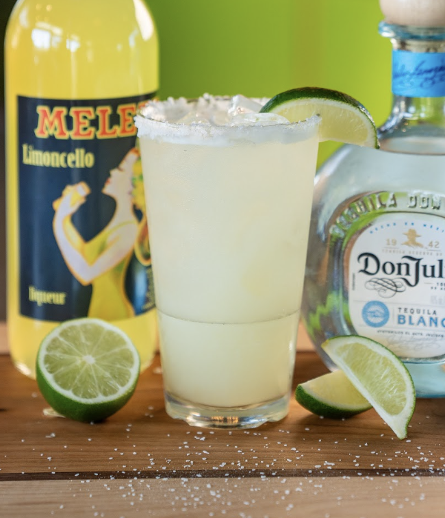 A margarita with a salted rim and lime wedge sits between a bottle of Don Julio tequila and a Mele Limoncello bottle, with lime halves and salt scattered on a wooden surface.