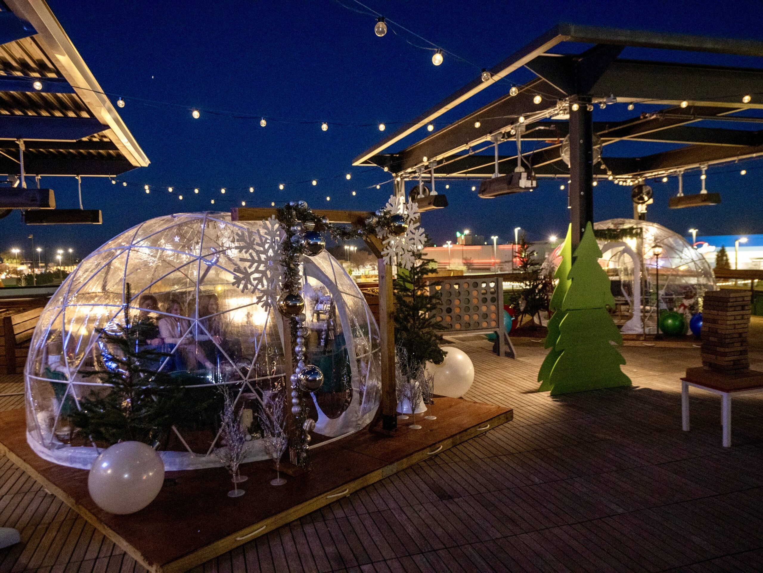 Outdoor rooftop with glowing string lights and clear igloo domes decorated for winter; people dine inside, surrounded by festive trees and ornaments at night.