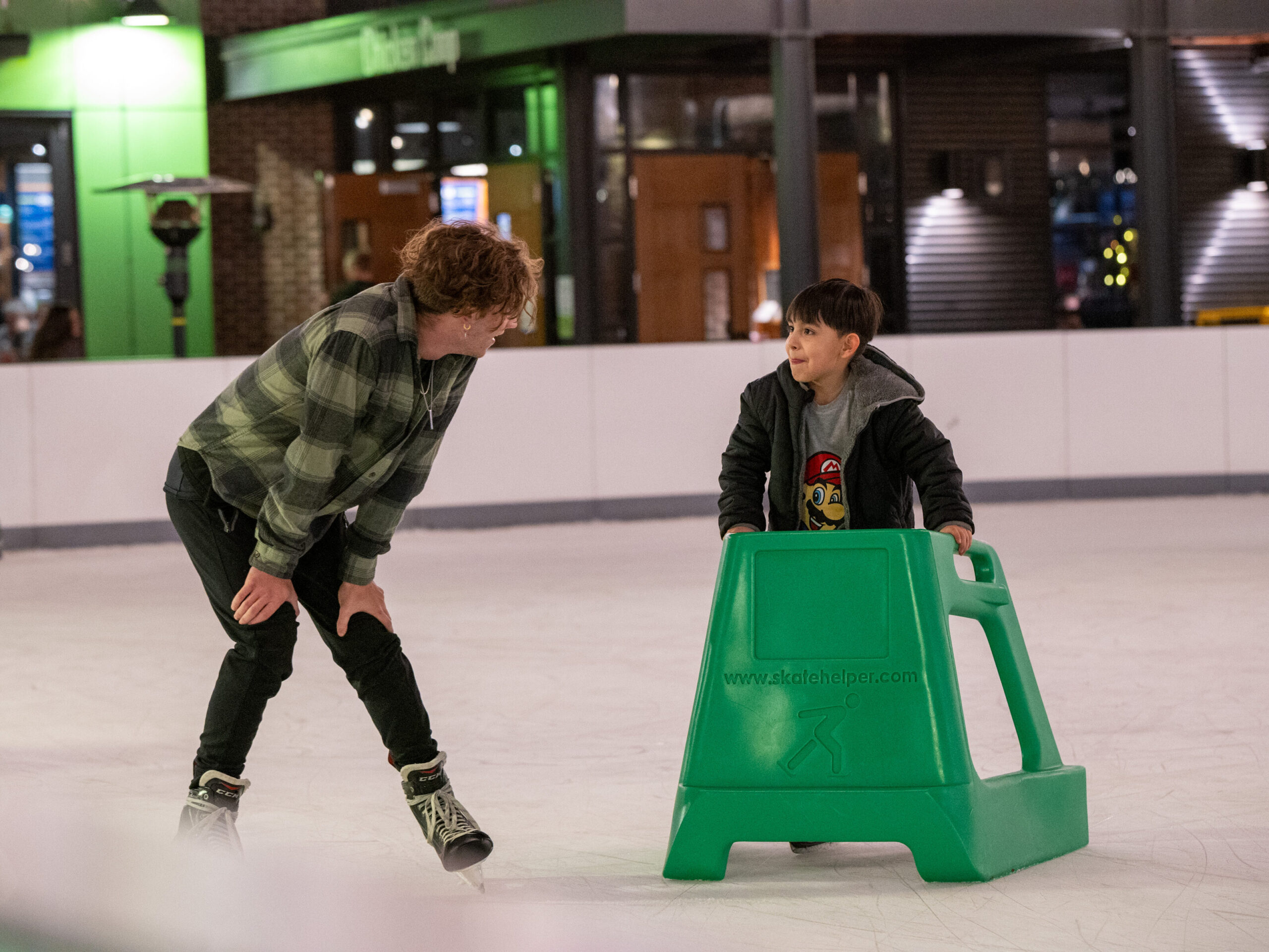 Two people ice skating indoors; an adult in a green plaid shirt skates beside a child using a green skate aid. Both are smiling and appear to be talking. Other skaters are visible in the background.
