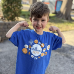 Young child smiling and flexing their arms, wearing a bright blue “Griffman’s Army” T-shirt with various colorful icons outside on a sunny day. Trees and a blurred background are visible.