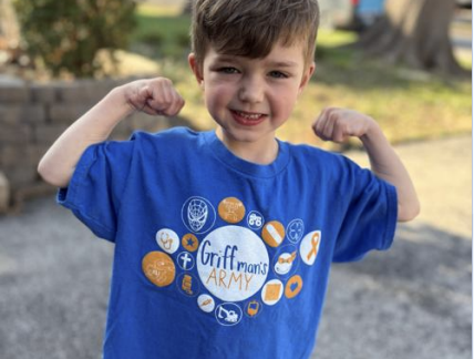Young child smiling and flexing their arms, wearing a bright blue “Griffman’s Army” T-shirt with various colorful icons outside on a sunny day. Trees and a blurred background are visible.