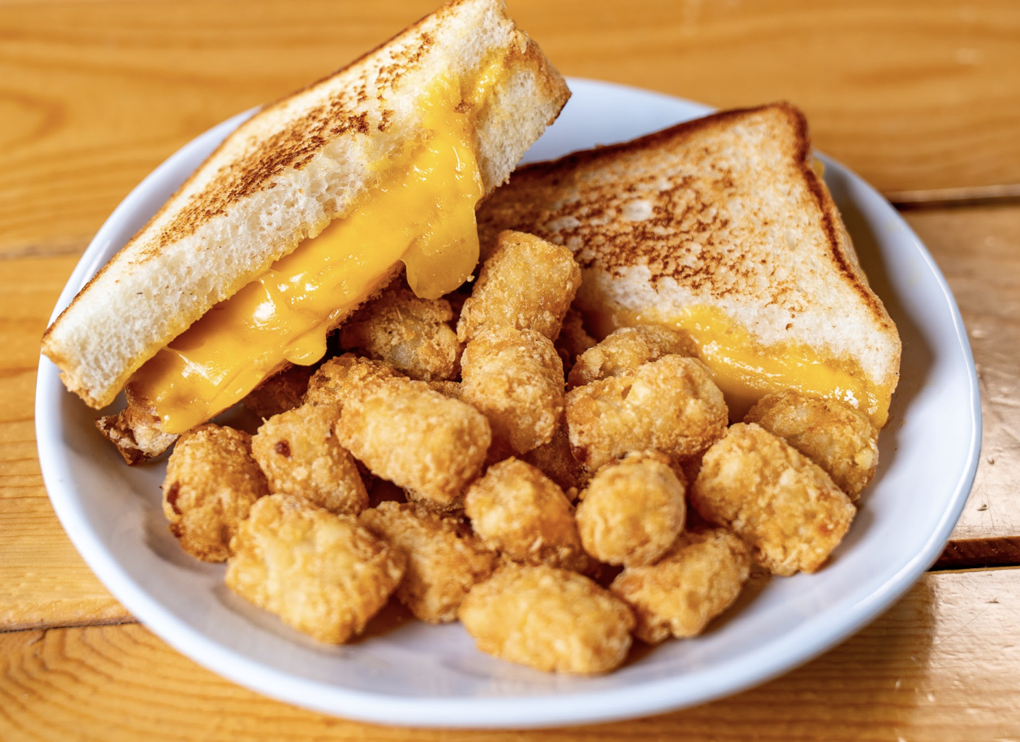 A white plate with a grilled cheese sandwich cut in half and filled with melted cheddar, served with a pile of crispy tater tots on a wooden table.