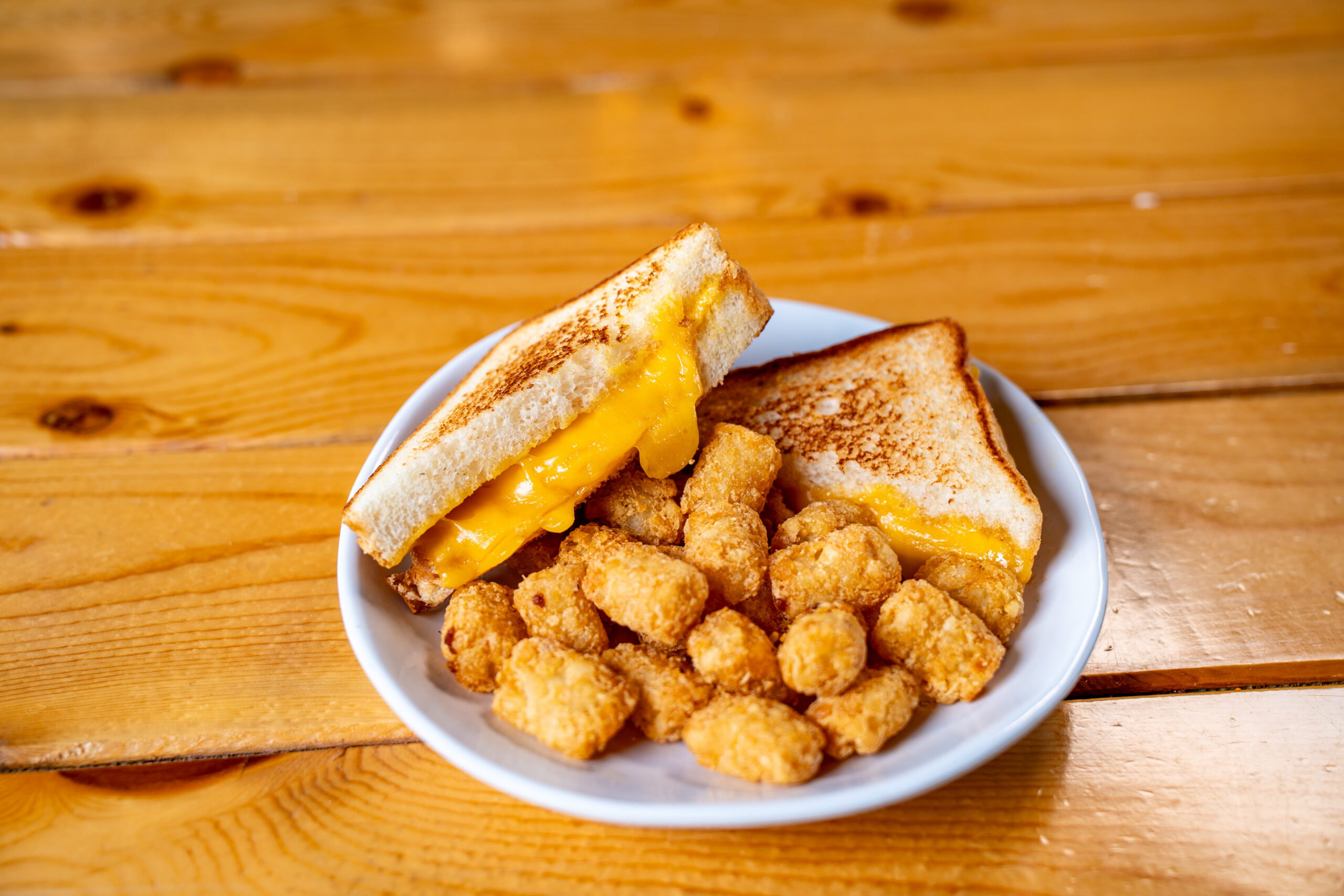 A plate with a grilled cheese sandwich cut in half, gooey cheddar cheese inside, and a serving of golden-brown tater tots, set on a wooden table.