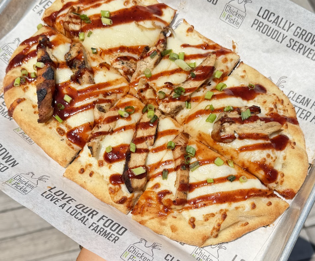 Flatbread pizza topped with melted cheese, grilled chicken, barbecue sauce drizzle, and chopped green onions, served on branded parchment paper on a metal tray.