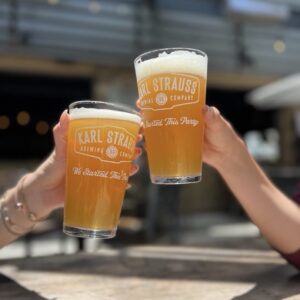 Two people clink glasses of beer labeled “Karl Strauss Brewing Company” at an outdoor table, with blurred background and sunlight highlighting the beverages.