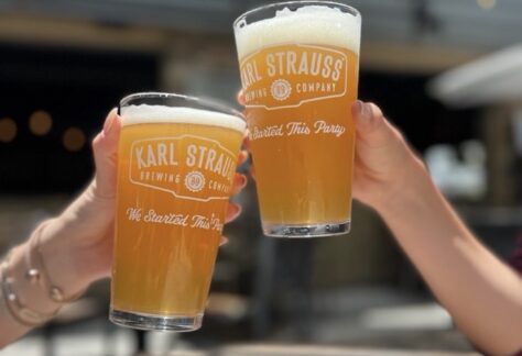 Two people clink glasses of beer labeled “Karl Strauss Brewing Company” at an outdoor table, with blurred background and sunlight highlighting the beverages.