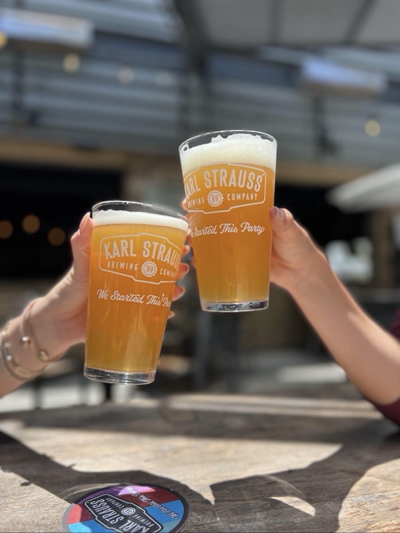 Two people clink glasses of beer labeled “Karl Strauss Brewing Company” at an outdoor table, with blurred background and sunlight highlighting the beverages.