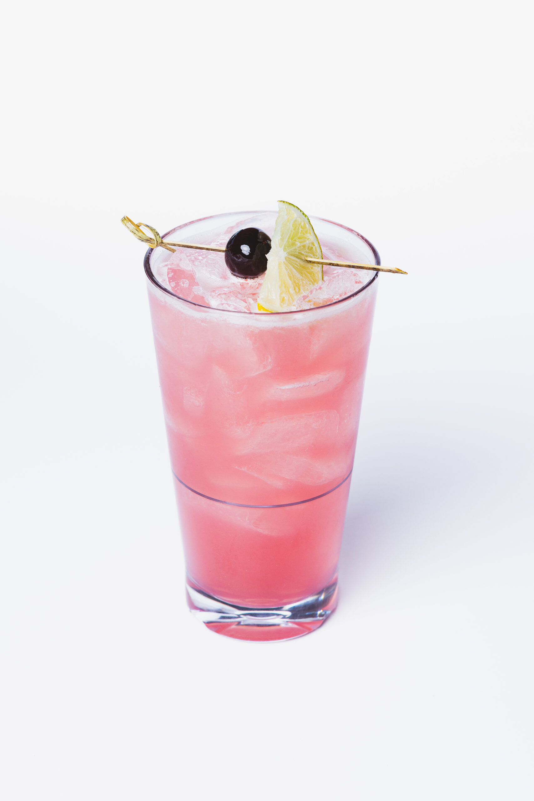 A pink iced cocktail in a clear glass, garnished with a lime wedge and a cherry on a cocktail pick, set against a white background.