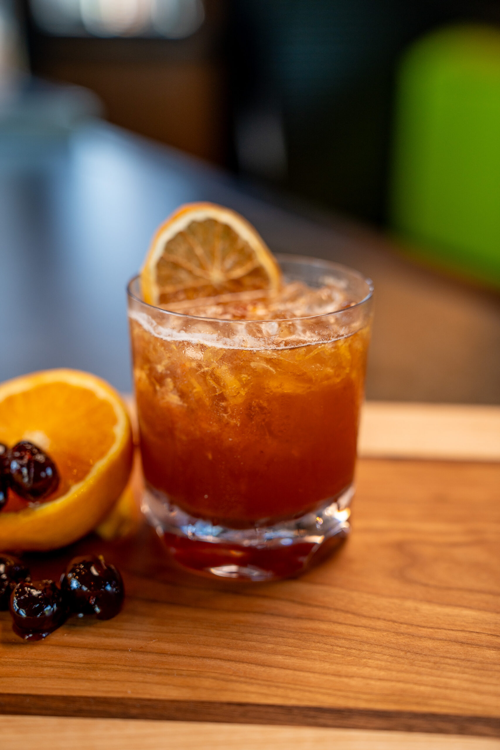 A cocktail in a glass with crushed ice, garnished with a dried orange slice. Beside it are half an orange and two dark cherries on a wooden surface.