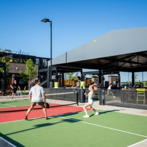 Four people play pickleball on an outdoor court under clear blue skies. A modern building with a covered patio and green accents is nearby. Spectators sit and watch the game.