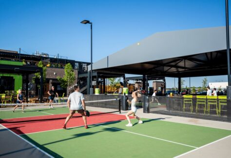 Four people play pickleball on an outdoor court under clear blue skies. A modern building with a covered patio and green accents is nearby. Spectators sit and watch the game.