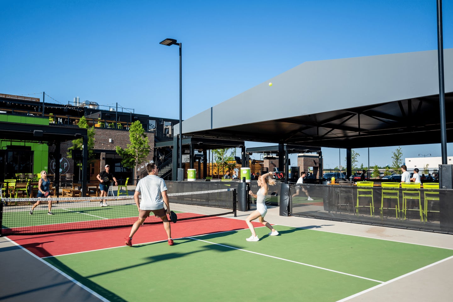 Four people play pickleball on an outdoor court under clear blue skies. A modern building with a covered patio and green accents is nearby. Spectators sit and watch the game.