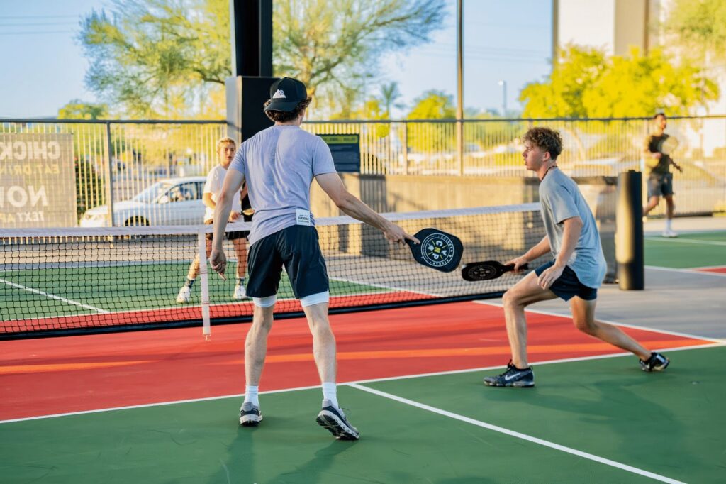 Two young men play pickleball on an outdoor court, holding paddles and facing each other near the net, with another person and greenery visible in the background.
