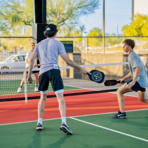 Two young men play pickleball on an outdoor court, holding paddles and facing each other near the net, with another person and greenery visible in the background.