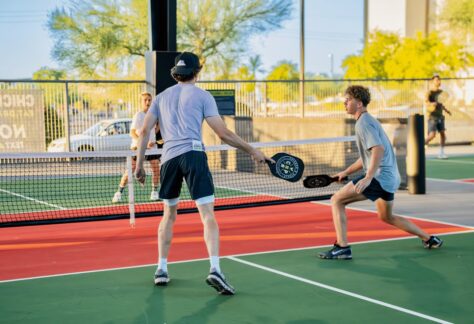 Two young men play pickleball on an outdoor court, holding paddles and facing each other near the net, with another person and greenery visible in the background.