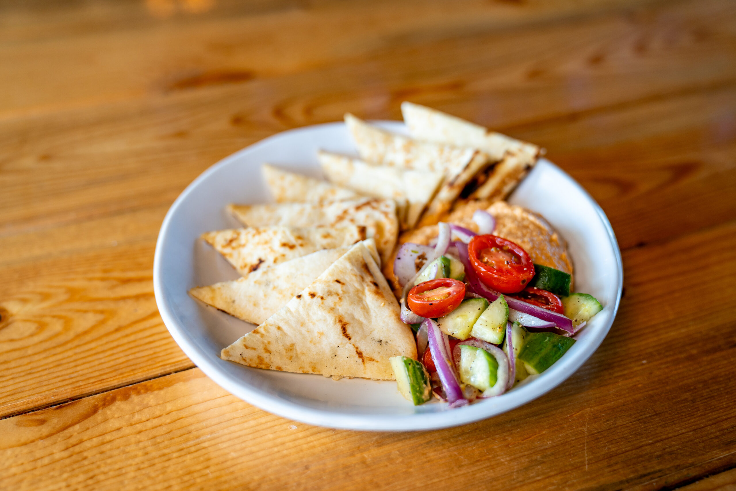 A white plate with pita bread slices, a serving of hummus, and a small salad with cucumber, cherry tomatoes, and red onion on a wooden table.