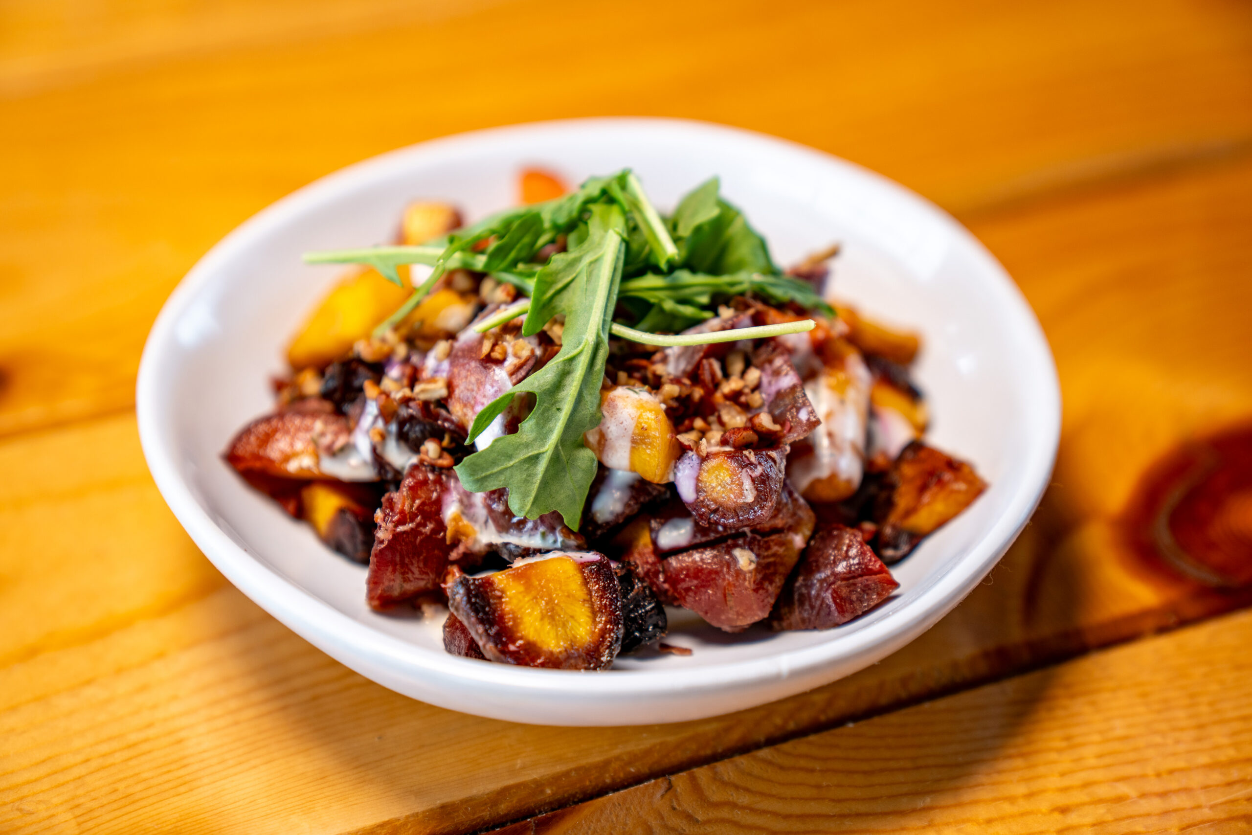 A white bowl holds a colorful salad with roasted sweet potatoes, greens, diced vegetables, and a creamy dressing, set on a wooden table.