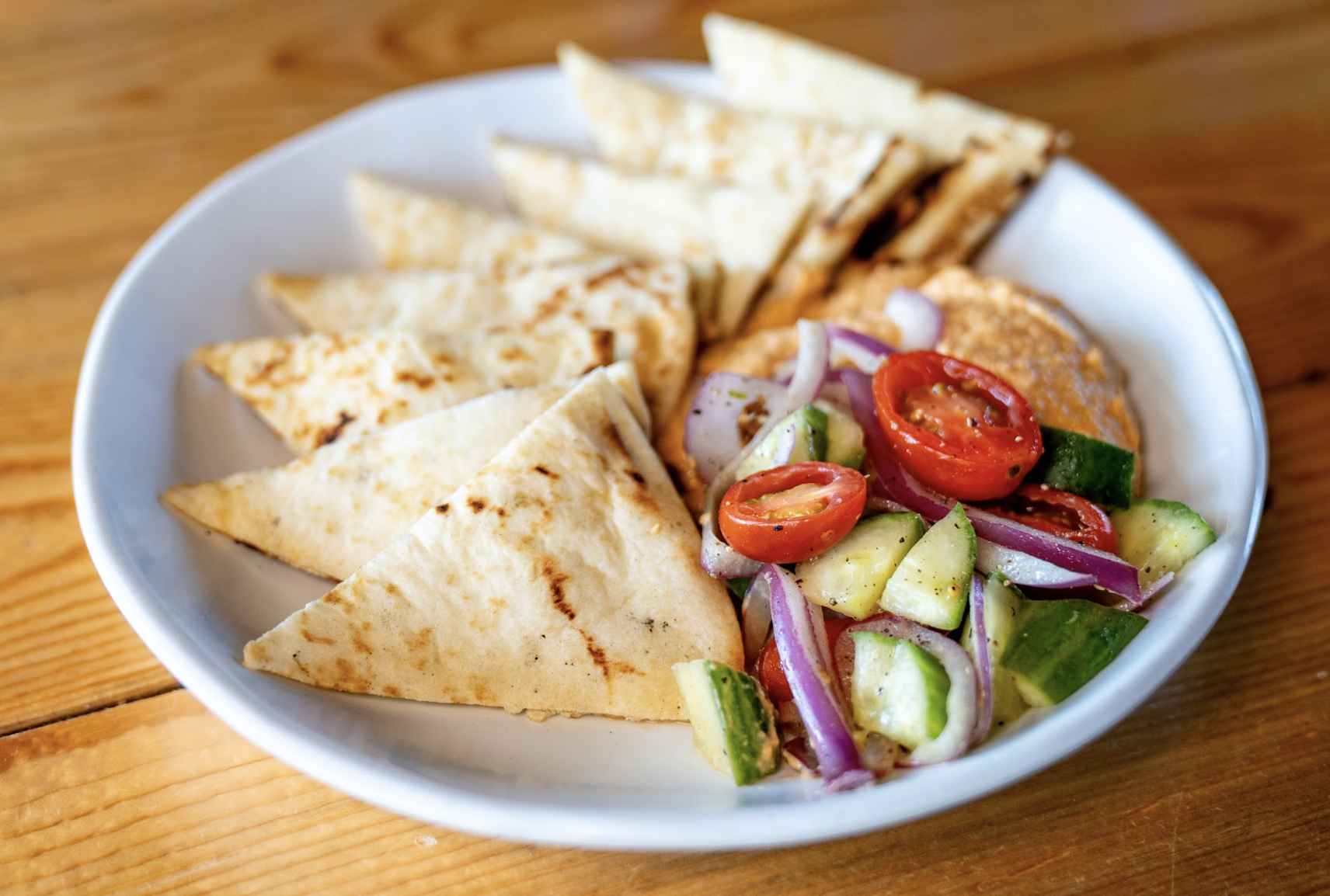 A white plate with pita triangles, hummus, and a salad of cherry tomatoes, cucumber, and red onion on a wooden table.