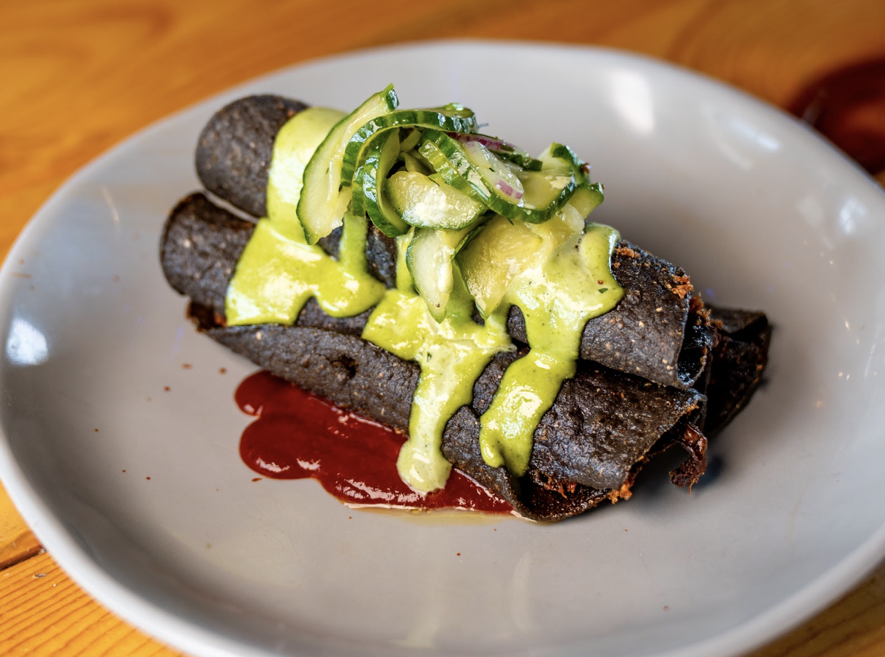 Three rolled blue corn tortillas on a white plate, topped with green sauce and sliced cucumbers, with a red sauce underneath.